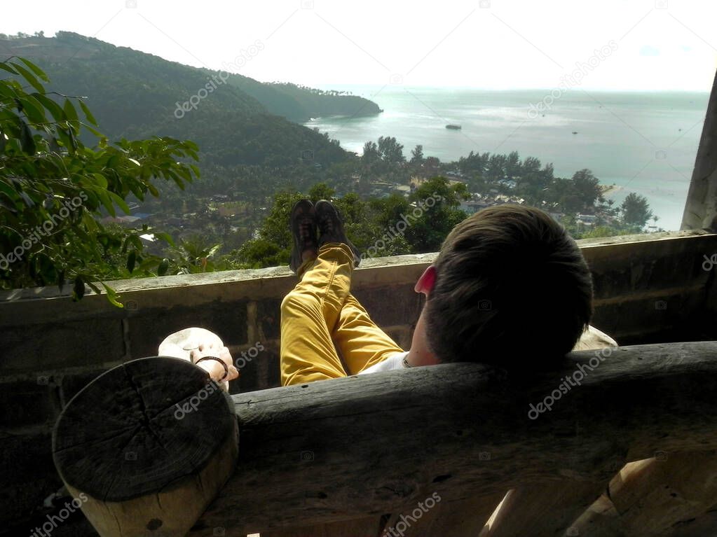 a man sits on a wooden chair admiring the view of the sea and mountains from a height.