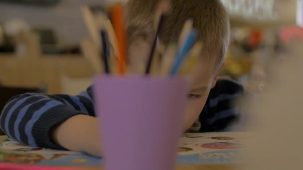 Liten pojke löser barnen träning i att utveckla bok för förskolebarn ofokuserat glas med pennor på förgrunden — Stockvideo