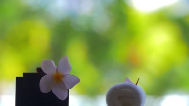 Vista en cámara lenta de pétalos de flores cayendo sobre fondo verde borroso sobre la mesa con flor Plumeria y toallas — Vídeos de Stock