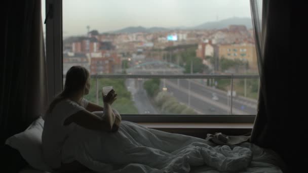 Woman in bed drinking tea and looking at city — Stock Video