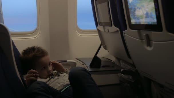 Вид маленького мальчика, смотрящего фильмы в гарнитуре в самолете, лежа на сиденье у окна самолета — стоковое видео