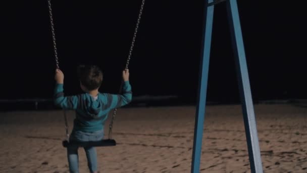 Boy swinging in the evening and looking at dark sea — Stock Video