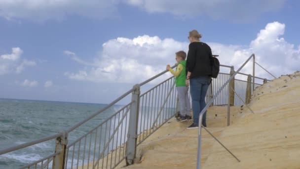 Mamá y el niño mirando al mar cuando salpicaduras altas que aparecen delante de ellos — Vídeos de Stock