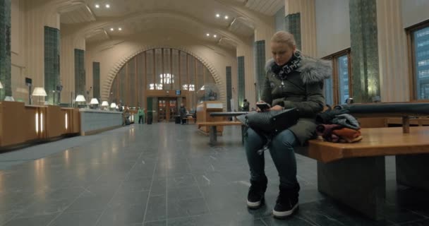 Kobieta czeka w hali dworca kolejowego i przy użyciu smartfona — Wideo stockowe