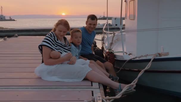 Föräldrar med äldste sonen och baby sitter på bryggan i solnedgången — Stockvideo