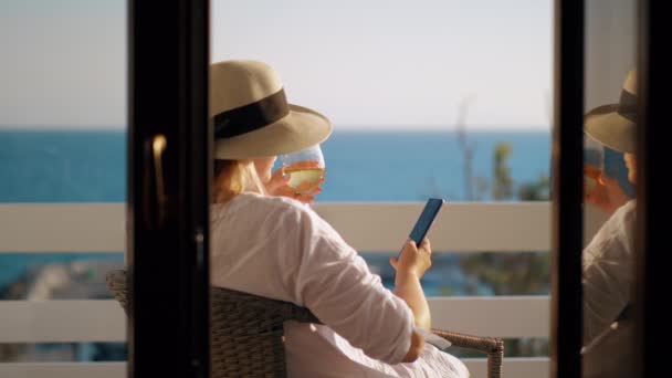 Mujer con copa de vino utilizando la celda cuando se relaja en el balcón con vistas al mar — Vídeo de stock