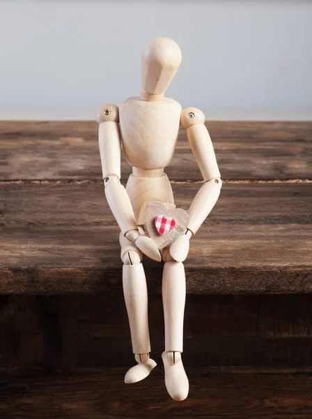 Wooden doll with heart in hands sitting on a wooden table. — Stock Photo, Image
