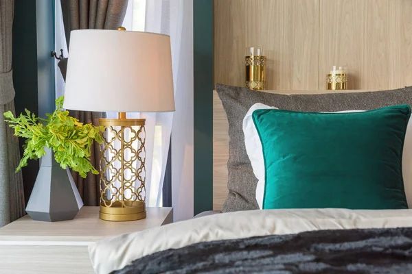 A gold luxury lamp on a beige table beside the bed with a fcandlestick on headboard and green pillow on a bed and curtain in bedroom with light from window.