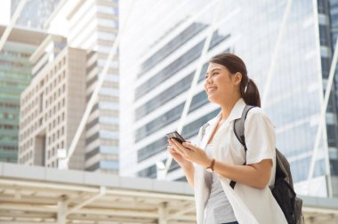 Düşük açılı, orta ölçekli, gülümseyen, sıradan beyaz tişörtlü Asyalı bir kadın sırt çantası taşıyor ve elinde bir cep telefonu tutuyor..