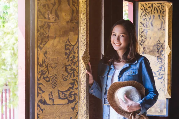 ミディアムクローズアップポートレート笑顔若いアジア人女性でジーンズジャケットとわら帽子立って次のタイの木のテクスチャウィンドウで寺院での旅行 — ストック写真