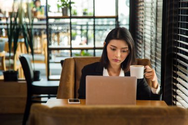 Orta boy. Resmi takım elbiseli çekici Asyalı kadın pencerenin yanındaki masada dizüstü bilgisayarla çalışıyor. Elinde bir fincan kahveyle sabah haberlerini kontrol ediyor ve çalışıyor..
