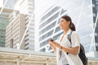 Düşük açılı, orta ölçekli, gülümseyen, sıradan beyaz tişörtlü Asyalı bir kadın sırt çantası taşıyor ve elinde bir cep telefonu tutuyor..