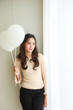 Orta boy, beyaz balon tutan ve yan panjurlara bakan bej tişörtlü güzel Asyalı kadın portresi..