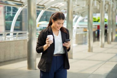 Resmi olarak gülümseyen genç Asyalı kadın, modern yaya köprüsünde yürürken elinde bir fincan kahveyle cep telefonunu seyrederek neredeyse ofis binasına doğru yürüyordu.