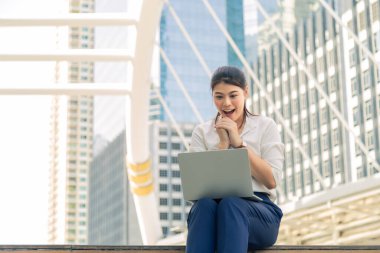 Modern yaya köprüsünde yerde oturan beyaz tişörtlü, gülümseyen Asyalı genç bir kadın, dizüstü bilgisayarından gelen güzel haberlerle heyecan verici.. 