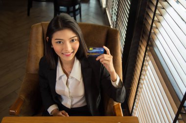 Orta ölçekli, yakışıklı, resmi takım elbiseli bir Asyalı kadın modern ofiste elinde bir kredi kartıyla oturuyor ve kameraya bakıyor.