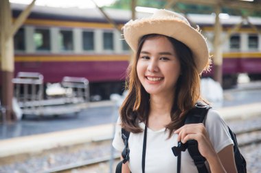 Orta yakın bir portre kot ceket ve hasır şapka giyen güzel Asyalı kadın sırtında sırt çantasıyla kamera tebessümüne bakıyor sıradaki tren yolunda mutlu bir şekilde seyahat ediyor..