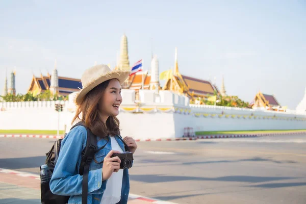 Medium close up portrait beautiful young Asian woman in jeans jacket and straw hat carrying a backpack holding a retro camera excited about traveling in Thailand in front of Wat Phra Kaew background.