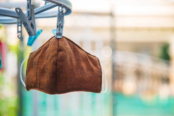 Kullanılmış kahverengi grip maskesi ön bahçedeki evin bulanık arka planıyla güneş ışığı altında plastik mandalla asılı duruyor..