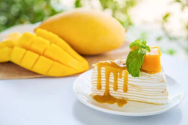 Mango krepli pasta beyaz tabakta nane şekeri ve krem şantili beyaz masada mango meyveli ve dilimlenmiş arka planda..