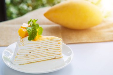 Mango meyveli ve yeşil doğa arkaplanlı bir nane şekeriyle süslenmiş beyaz bir tabağa dilimlenmiş mango krepi ve nane şekerini kapatın..