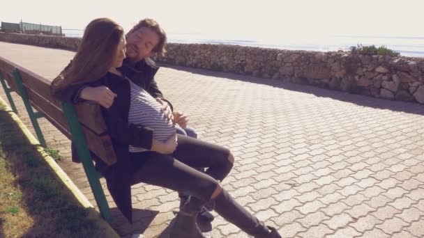 Ζευγάρι στην αγάπη ευτυχής χαμογελαστοί κάθεται σε παγκάκι, μπροστά στον ωκεανό άνδρα να κρατάει την κοιλιά εγκύου — Αρχείο Βίντεο