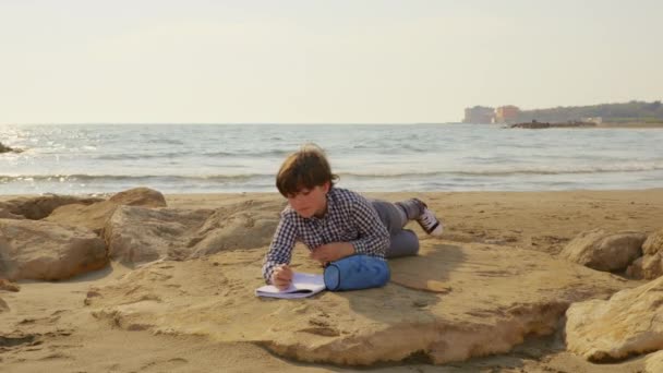 Παιδί που κάνει την εργασία μπροστά από τον ωκεανό στο ηλιοβασίλεμα. — Αρχείο Βίντεο