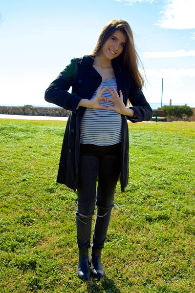 Preciosa mujer embarazada mostrando signo de amor en el vientre en el parque de tiro ancho —  Fotos de Stock