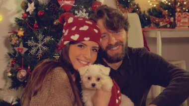 Mutlu çift Noel gecesi, küçük yavru köpeğe sarılıyor ve kameraya bakıyor.