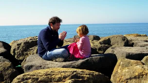 Feliz niña jugando con el padre sentado en las rocas frente al océano 4K cámara lenta — Vídeo de stock