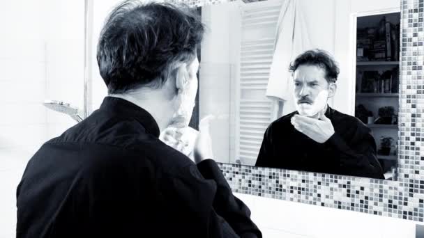 Κομψό όμορφος άντρας βάζει κρέμα ξυρίσματος στο πρόσωπο μπροστά από καθρέφτη στο σπίτι άσπρο και μαύρο — Αρχείο Βίντεο