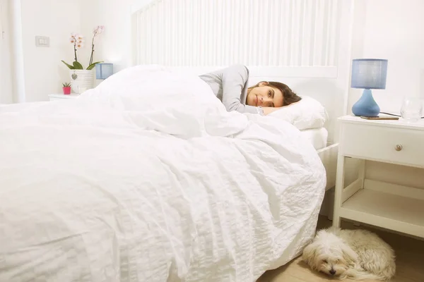 Mujer acostada en la cama junto a un perro feliz mirando la cámara — Foto de Stock