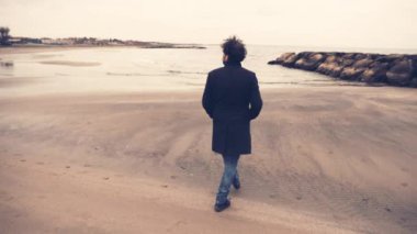 Kışın paltolu şık yakışıklı bir adam okyanusun önünde yürüyor ve düşünüyor.