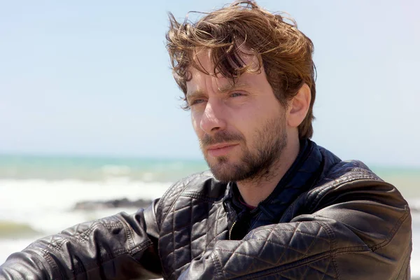 Handsome man with blue eyes and beard sitting in front of ocean in winter looking view closeup — Stock Photo, Image