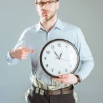 Pensativo hombre de negocios señalando en reloj aislado en gris