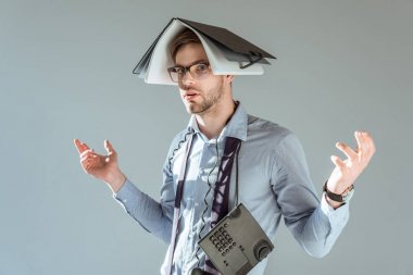 Young confused businessman holds notebook on his head and phone on neck isolated on grey clipart