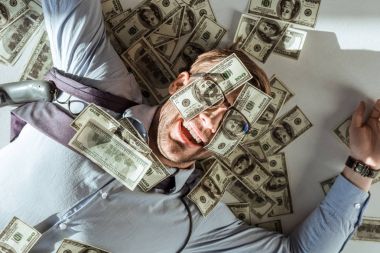 Young happy rich businessman lying on floor with dollar bills covering his face