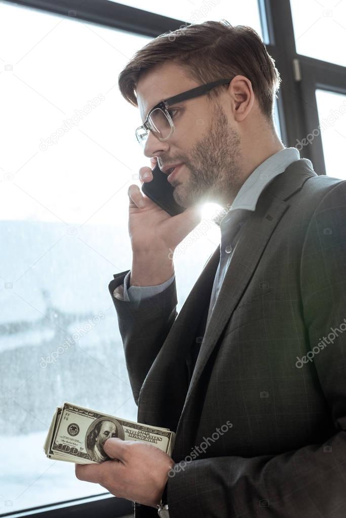 Young confident businessman holding money and talking on the smartphone by the window
