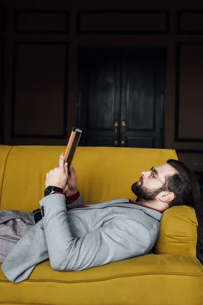 躺在黄色沙发上用数码片的大胡子男子 — 图库照片