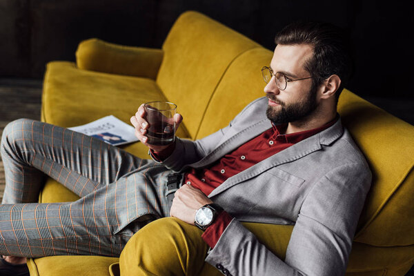 elegant man drinking whiskey and sitting on sofa with newspaper 