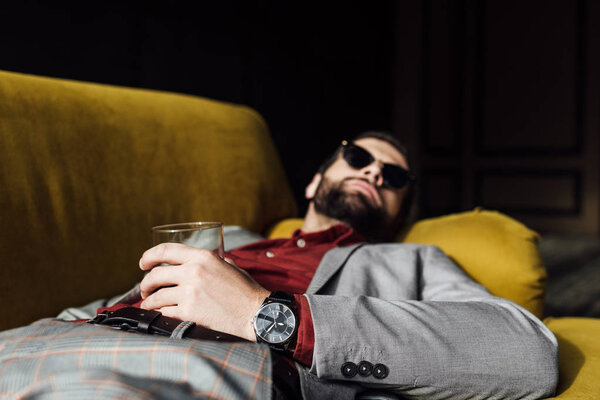 drunk man in sunglasses with glass of cognac lying on couch