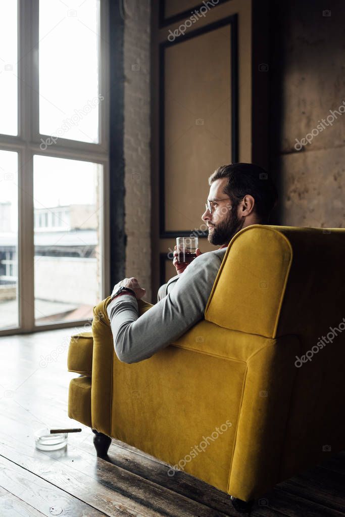 stylish handsome man holding glass of whiskey and sitting on sofa