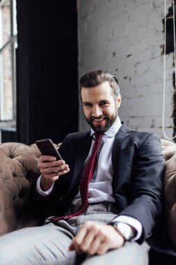 handsome smiling businessman using smartphone and sitting in armchair  clipart
