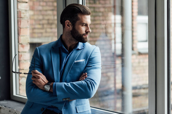 задумчивый мужчина в синем модном костюме держит очки и смотрит в окно
