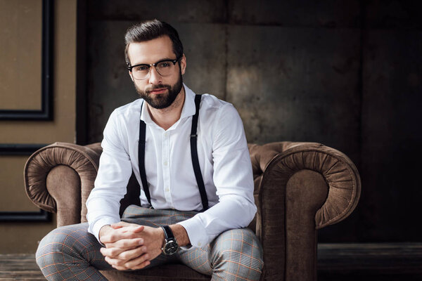 handsome stylish man in glasses and suspenders sitting in armchair 