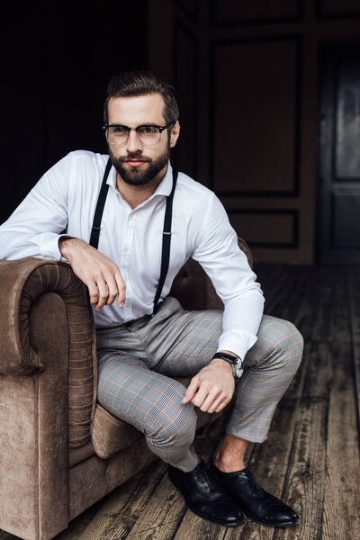 handsome bearded man in eyeglasses and suspenders sitting in armchair 