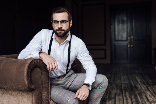 fashionable bearded man in glasses and suspenders sitting in armchair 