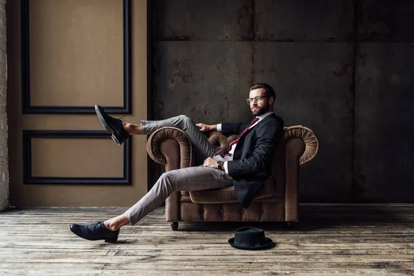 Przystojny Biznesmen Elegancki Pozowanie Fotel Kapelusz Leżąc Podłodze Pobliżu Wnętrze — Zdjęcie stockowe