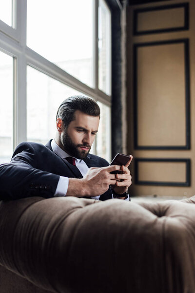 fashionable bearded businessman messaging on smartphone while sitting in armchair 