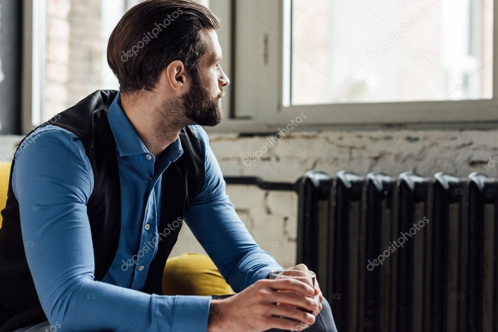 elegant bearded man holding alcohol drink and sitting on yellow sofa